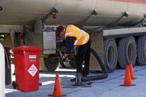 A worker in a high-visibility vest connecting a large hose to a tanker truck next to a red spill containment bin.